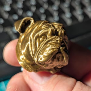 Puede incluir: Una cabeza de bulldog dorada, impresa en 3D. La escultura tiene arrugas y pliegues detallados, con la boca ligeramente abierta y papada visible. Las orejas del bulldog están levantadas y el diseño es muy texturizado.