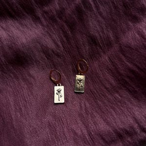 May include: A pair of gold-colored earrings. Each earring features a rectangular pendant with a floral design. The earrings are displayed against a dark purple, textured fabric background.