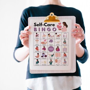 May include: A white clipboard with a purple and pink self-care bingo card. The card has 25 squares with different self-care activities illustrated in each square. The title of the card is "Self-Care Bingo".