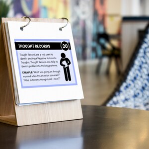Puede incluir: Una tarjeta blanca con texto azul y negro en un soporte de madera. La tarjeta est&aacute; titulada "Thought Records" e incluye un ejemplo de c&oacute;mo usar los registros de pensamientos. La tarjeta est&aacute; numerada 20.