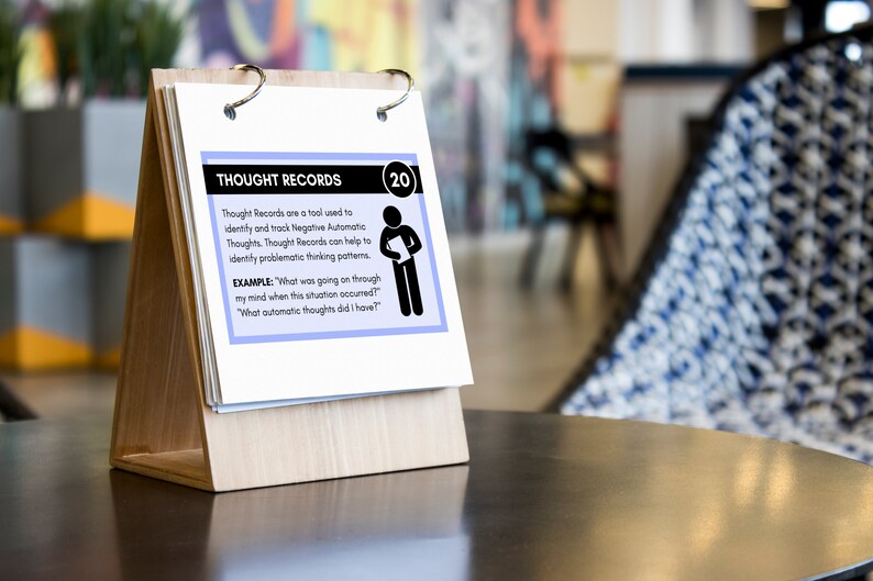 Puede incluir: Una tarjeta blanca con texto azul y negro en un soporte de madera. La tarjeta est&aacute; titulada "Thought Records" e incluye un ejemplo de c&oacute;mo usar los registros de pensamientos. La tarjeta est&aacute; numerada 20.