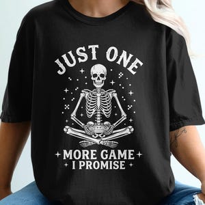 May include: Black t-shirt with a white skeleton graphic. The skeleton is seated, holding a game controller, with the text "JUST ONE + MORE GAME + I PROMISE +" above and below the image. The shirt is a casual wear item.