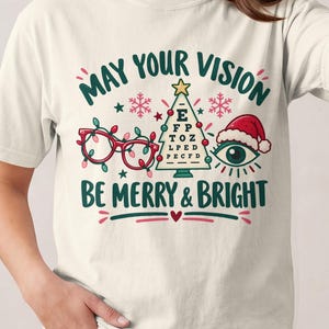 May include: A cream-coloured t-shirt featuring a graphic with the text "MAY YOUR VISION BE MERRY & BRIGHT". The design includes red spectacles wrapped in Christmas lights, a green eye chart shaped like a Christmas tree, and an eye wearing a Santa hat.