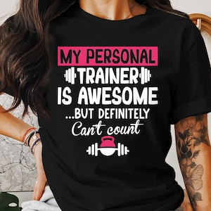 Pode incluir: Camiseta preta com texto branco que diz "My Personal Trainer is Awesome...but definitely can't count". O texto é emoldurado por retângulos rosa. Um kettlebell rosa é representado abaixo do texto.