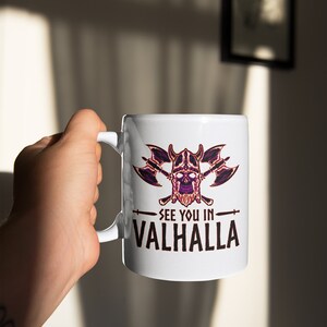 Puede incluir: Taza de cer&aacute;mica blanca con un dise&ntilde;o de calavera negro y morado con hachas cruzadas y el texto "See you in Valhalla".