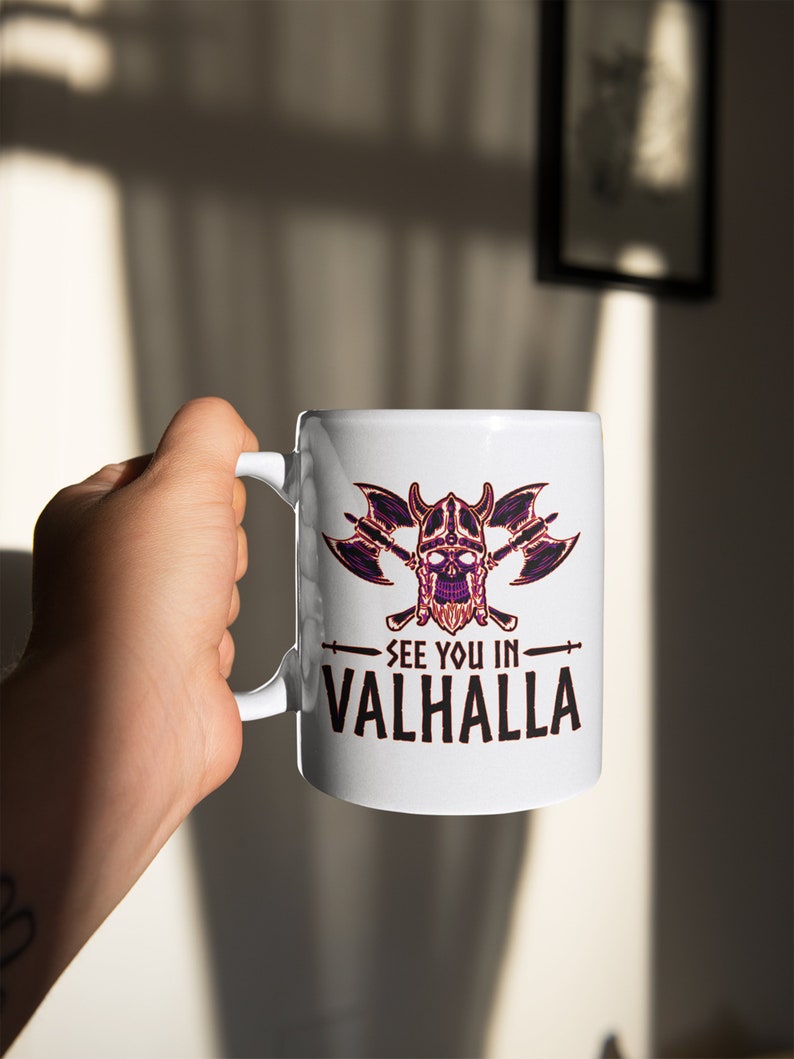 Puede incluir: Taza de cer&aacute;mica blanca con un dise&ntilde;o de calavera negro y morado con hachas cruzadas y el texto "See you in Valhalla".