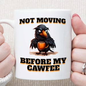 May include: White ceramic mug featuring a black cartoon crow holding a coffee cup. The mug has the text "NOT MOVING BEFORE MY CAWFFEE" in black and yellow.