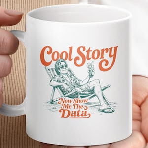 May include: A white ceramic mug featuring a graphic illustration of a person relaxing in a beach chair while holding a drink. The text "Cool Story" appears in orange above the illustration, and "Now Show Me The Data" is printed in orange below.