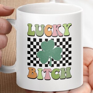 May include: White ceramic mug with a checkered background and a green four-leaf clover. The text "LUCKY BITCH" is written in colorful letters.