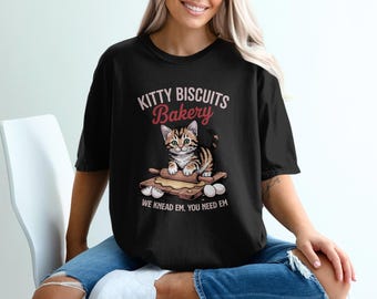 May include: Black t-shirt with a graphic of a kitten on a wooden board with a rolling pin and eggs. The text "Kitty Biscuits Bakery" is above the kitten, and "We knead em. You need em" is below.
