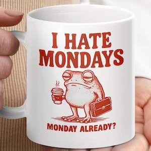May include: White ceramic mug featuring the red text "I HATE MONDAYS" and a cartoon frog with a coffee cup and briefcase. The mug also has the phrase "MONDAY ALREADY?" printed on it.