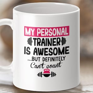 May include: White ceramic coffee mug with a pink and black design. The text on the mug reads "My Personal Trainer Is Awesome...But Definitely Can't Count".