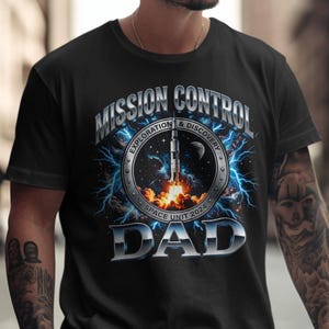 May include: Black t-shirt with a space-themed graphic. The design features the words "MISSION CONTROL DAD" in silver, a rocket launching, and the text "EXPLORATION & DISCOVERY SPACE UNIT 2024". Lightning bolts and a starry background complete the design.