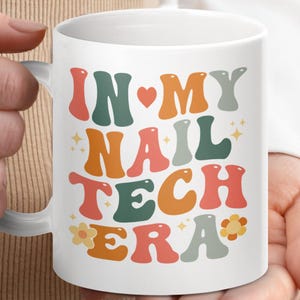 May include: White ceramic mug with a colourful retro design that says "In My Nail Tech Era" with a heart and flowers.