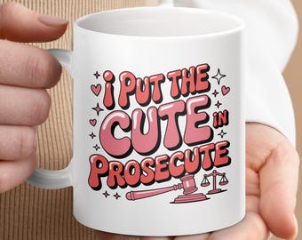 May include: White ceramic mug with the text "I PUT THE CUTE IN PROSECUTE" in pink and red bubble letters. The mug also features a gavel, scales of justice, hearts, and stars.