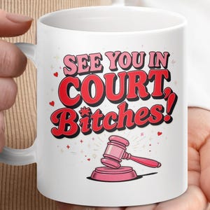 May include: White ceramic mug with the text "SEE YOU IN COURT Bitches!" in red and pink lettering. A pink gavel and sound block are below the text. The mug is held by a person.