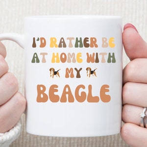 May include: White ceramic mug with the text "I'd rather be at home with my beagle" in colourful, retro-style lettering. Two beagle dog illustrations are included in the text.