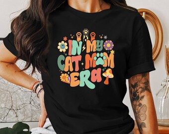 May include: Black t-shirt with a colorful graphic that says "In My Cat Mom Era" with flowers, mushrooms, and paw prints.