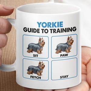 Puede incluir: Taza de cerámica blanca con una Guía de Entrenamiento para Yorkies. La taza muestra ilustraciones de un Yorkshire Terrier demostrando los comandos: Sit, Paw, Fetch y Stay. El texto es azul y negro.