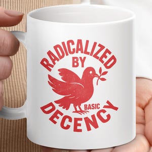 May include: White ceramic mug featuring a red dove graphic with an olive branch, and the text "RADICALIZED BY BASIC DECENCY". The mug has a curved handle. The mug is held by a person.