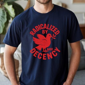 May include: Navy blue t-shirt featuring a red dove graphic with an olive branch and the text "RADICALIZED BY BASIC DECENCY". The design is in a distressed font. The t-shirt has a crew neck.