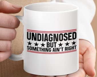 May include: White ceramic coffee mug with a red, white, and blue design. The text on the mug reads "Undiagnosed but something ain't right".