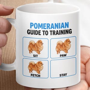 Puede incluir: Taza de cerámica blanca con una guía de entrenamiento para perros Pomerania. La taza tiene el texto "Pomeranian Guide to Training" en azul, con ilustraciones del perro demostrando "Sit", "Paw", "Fetch" y "Stay".