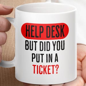 May include: White ceramic mug featuring the text "HELP DESK BUT DID YOU PUT IN A TICKET?" in black and red. The words "HELP DESK" are in a red oval. The mug is held by a person.