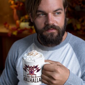 Puede incluir: Una taza de cer&aacute;mica blanca con un gr&aacute;fico negro y morado de una calavera con hachas cruzadas. La taza tiene el texto "See you in Valhalla" impreso en ella. La taza est&aacute; llena de una bebida cremosa cubierta con canela.