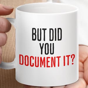 May include: White ceramic mug with the text "BUT DID YOU DOCUMENT IT?" in black and red lettering. The mug has a white handle and is held by a person.