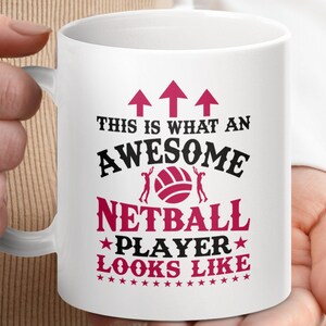 May include: White ceramic mug with a handle, featuring the text "THIS IS WHAT AN AWESOME NETBALL PLAYER LOOKS LIKE" in black and pink. The design includes arrows, a netball, and stars.