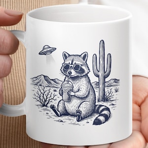 May include: White ceramic mug with a design of a raccoon in sunglasses, sipping from a cup with a straw. The illustration includes a desert scene with a cactus, mountains, and a UFO. The raccoon has a striped tail.