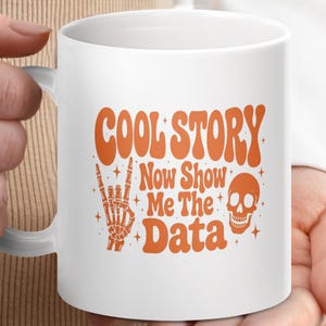 May include: A white ceramic mug featuring orange text that reads COOL STORY Now Show Me The Data. The design includes an orange graphic of a skeleton hand making a rock and roll gesture and an orange skull, accented with small four-pointed stars.