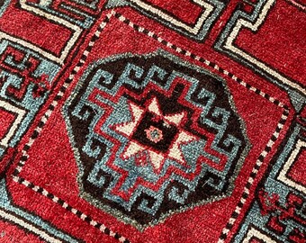 Puede incluir: Una alfombra roja con un patrón geométrico azul y blanco. La alfombra tiene un diseño tradicional con detalles intrincados.