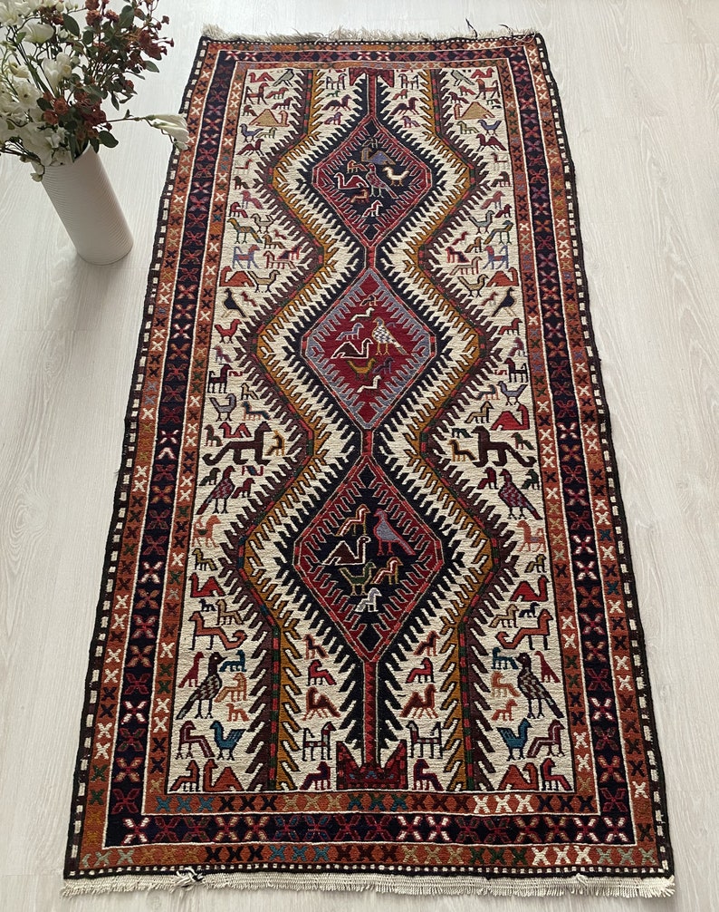 May include: A traditional rug with a cream background and a geometric pattern in shades of red, orange, blue, and black. The pattern features stylized birds and animals.