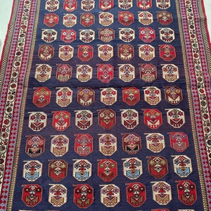 Op de afbeelding: Een traditioneel Turks tapijt met een donkerblauwe achtergrond en een herhalend patroon van rode, witte en gouden achthoeken. Het tapijt heeft een decoratieve rand met bloem- en geometrische motieven.