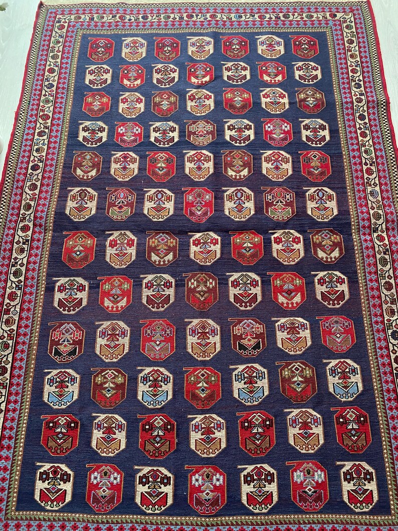Op de afbeelding: Een traditioneel Turks tapijt met een donkerblauwe achtergrond en een herhalend patroon van rode, witte en gouden achthoeken. Het tapijt heeft een decoratieve rand met bloem- en geometrische motieven.