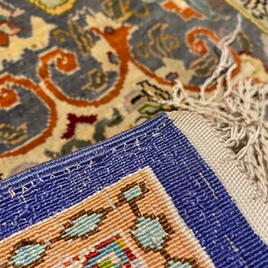 May include: Close-up of a blue and white rug with a detailed floral pattern. The rug has a fringe border and is woven with intricate details.