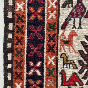 May include: Close-up of a colorful woven rug with intricate patterns. The rug features a border with geometric designs and a central field with stylized animal figures. The colors include red, orange, white, black, and green.