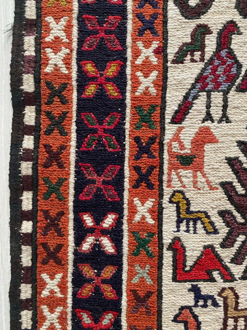 May include: Close-up of a colorful woven rug with intricate patterns. The rug features a border with geometric designs and a central field with stylized animal figures. The colors include red, orange, white, black, and green.