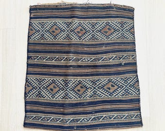 May include: A rectangular, woven textile with a geometric pattern. The design features alternating bands of dark blue and brown, with diamond and zigzag motifs in white and tan. The textile has a slightly worn appearance.
