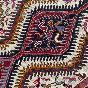May include: A close-up of a colorful woven rug with a geometric pattern. The rug features a central diamond shape with intricate designs in shades of red, blue, green, and black. The background of the rug is white with a repeating pattern of zigzags.