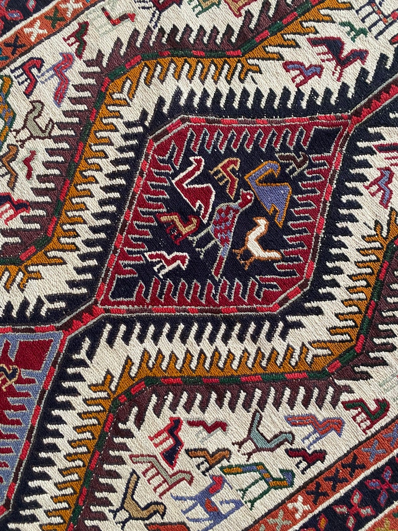 May include: A close-up of a colorful woven rug with a geometric pattern. The rug features a central diamond shape with intricate designs in shades of red, blue, green, and black. The background of the rug is white with a repeating pattern of zigzags.