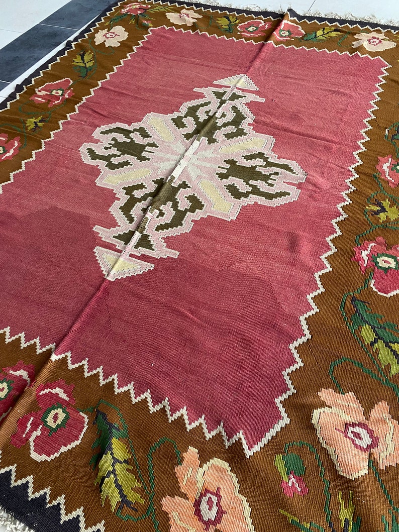 May include: A traditional style rug with a pink center and a brown border. The border features a floral pattern with pink, white, and green flowers.