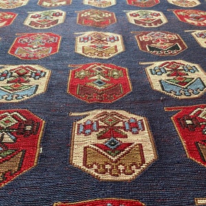 Op de afbeelding: Een close-up van een blauw tapijt met een herhalend patroon van rode, witte en bruine geometrische vormen. Het tapijt heeft een traditioneel ontwerp en is geweven met ingewikkelde details.