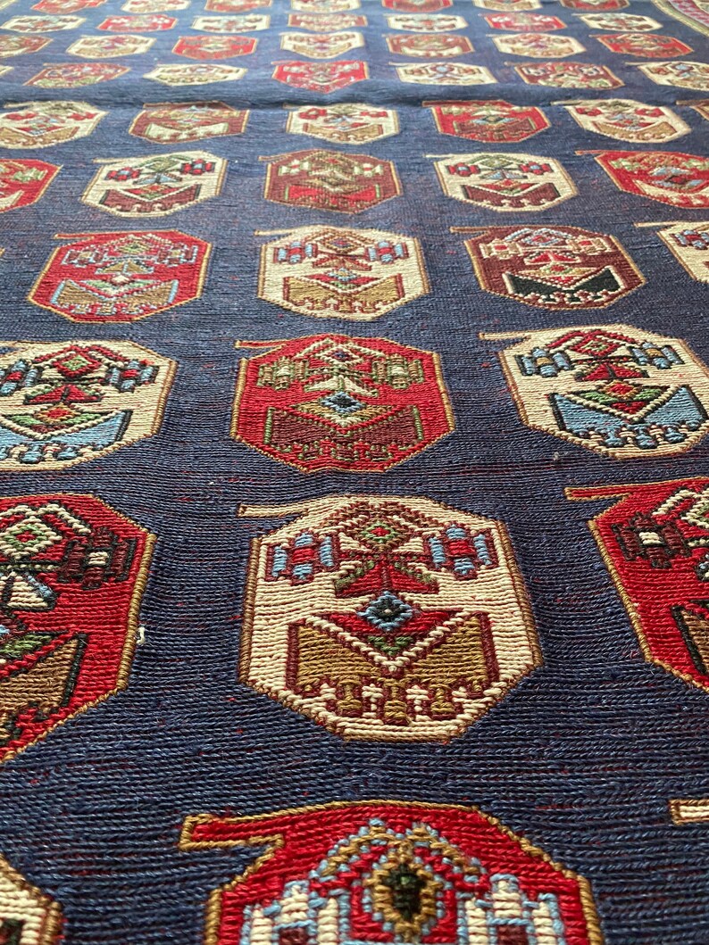Op de afbeelding: Een close-up van een blauw tapijt met een herhalend patroon van rode, witte en bruine geometrische vormen. Het tapijt heeft een traditioneel ontwerp en is geweven met ingewikkelde details.