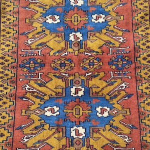 May include: A traditional rug with a red background and intricate geometric patterns in shades of blue, yellow, and brown. The rug features a central medallion with a repeating design.