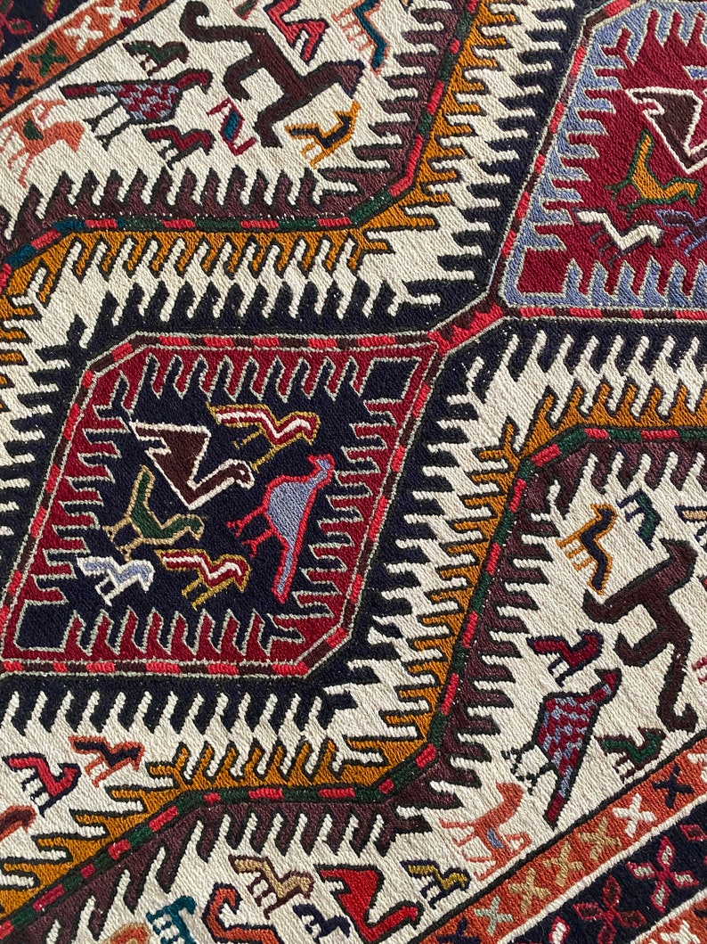 May include: Close-up of a colorful woven rug with geometric patterns and stylized animal figures. The rug features a white background with black, red, brown, blue, and yellow accents.