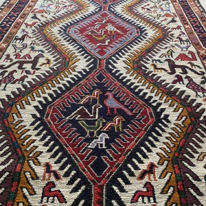 May include: A traditional rug with a geometric pattern in shades of red, brown, white, and gold. The rug features a central diamond shape with stylized birds and animals.