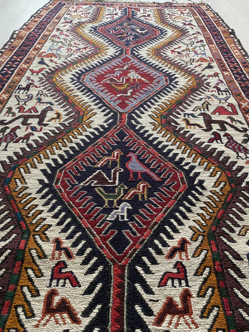May include: A traditional rug with a geometric pattern in shades of red, brown, white, and gold. The rug features a central diamond shape with stylized birds and animals.